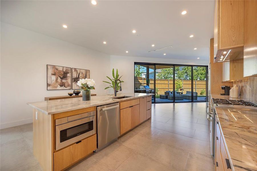 Kitchen featuring light stone countertops, light brown cabinetry, stainless steel appliances, decorative backsplash, and a center island with sink