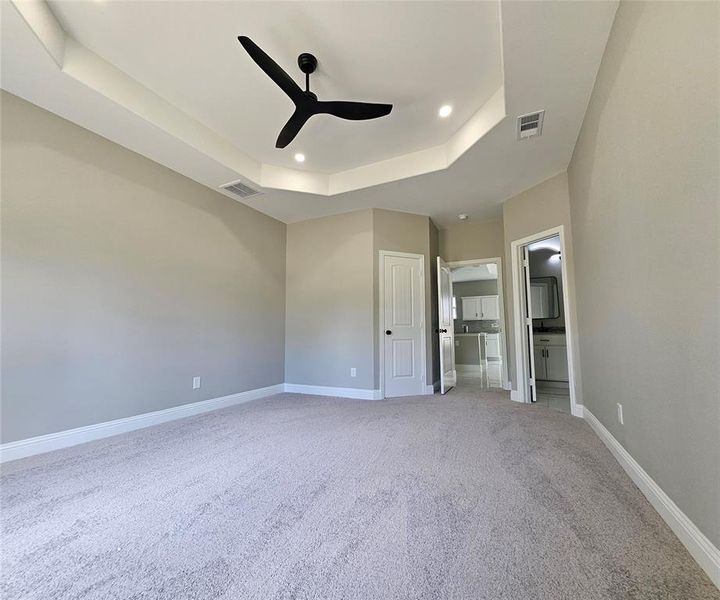 Master bedroom with ceiling fan, a raised ceiling, light carpet, and ensuite bath