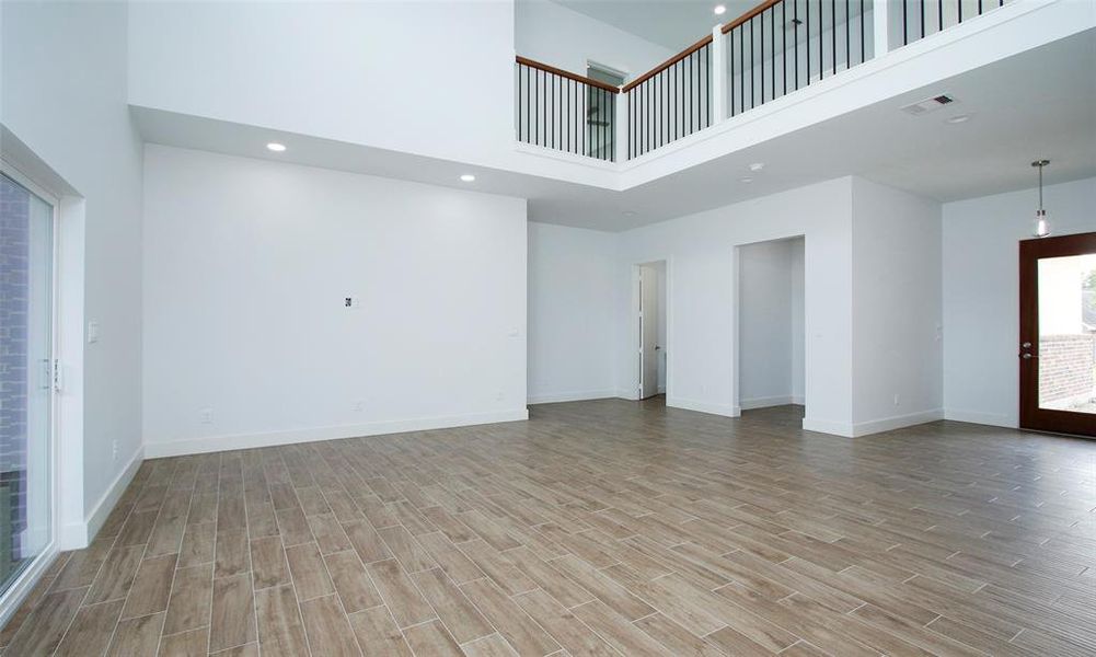 This is a modern, spacious kitchen with gray cabinetry, stainless steel appliances, granite countertops, and floating wooden shelves. It features a central island with a sink, contemporary pendant lighting, and large windows that provide ample natural light. The flooring is a wood-look tile, offering both durability and style.