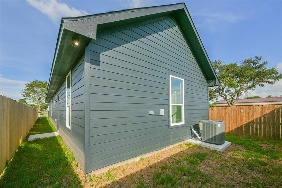 This photo shows the exterior side view of a modern home with blue-gray siding, featuring a well-maintained fence, a new HVAC unit, and a clear path along the side yard, all under a bright, sunny sky.