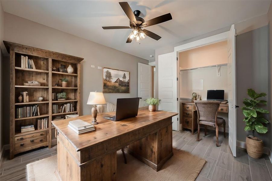 This is a cozy, well-appointed home office featuring a large wooden desk, a bookshelf filled with various decorative items and books, and a comfortable wicker chair. The room is finished with a neutral color palette, a ceiling fan, and a tasteful landscape painting, creating a tranquil workspace. There's also a built-in desk area in the nook, offering additional work space.