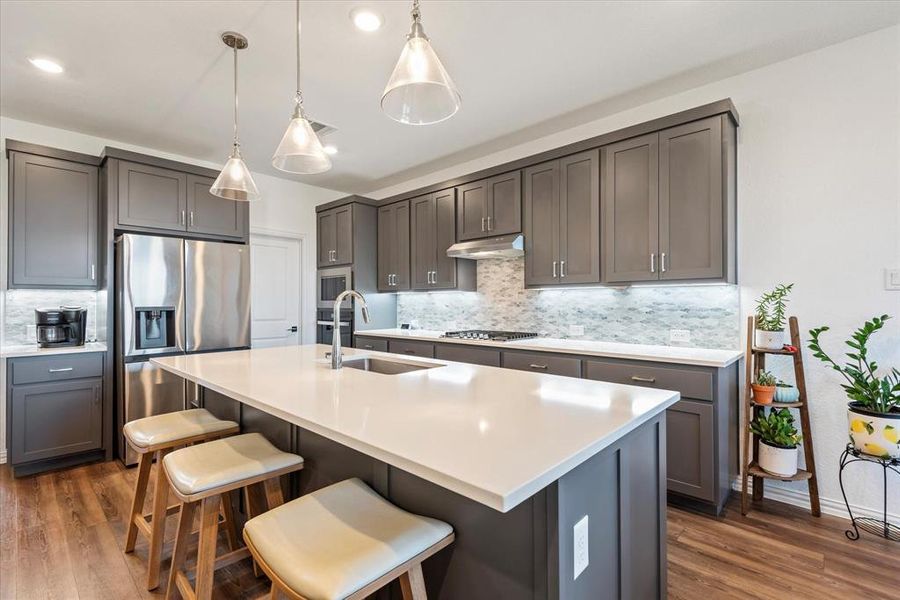 Kitchen with backsplash, stainless steel appliances, decorative light fixtures, dark wood-type flooring, and a kitchen island with sink