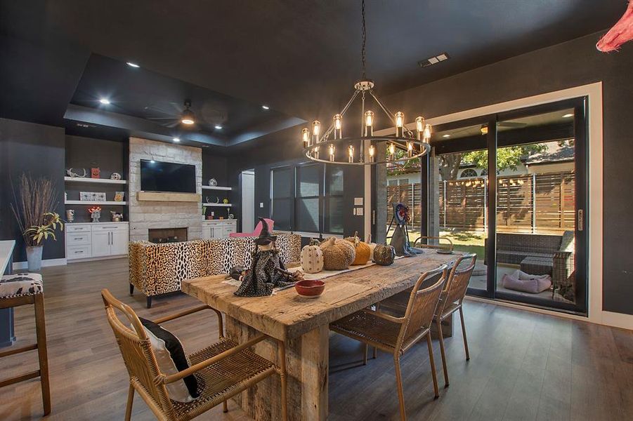 Dining space featuring a raised ceiling, dark wood-type flooring, ceiling fan with notable chandelier, built in shelves, and a stone fireplace