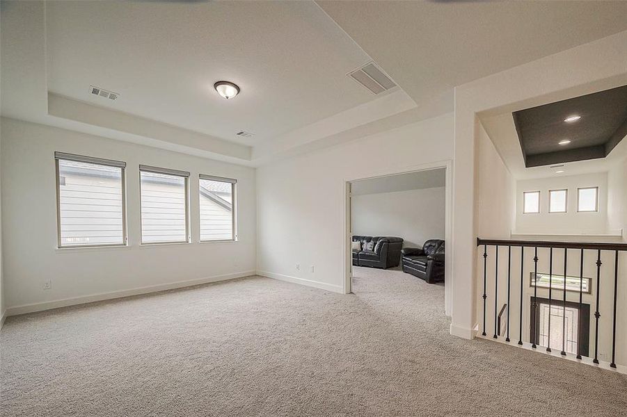 This is a spacious, carpeted room with high ceilings, recessed lighting, and three windows providing natural light. It features an open layout with a view of an adjacent room with a tray ceiling and is complemented by an elegant railing along the staircase.