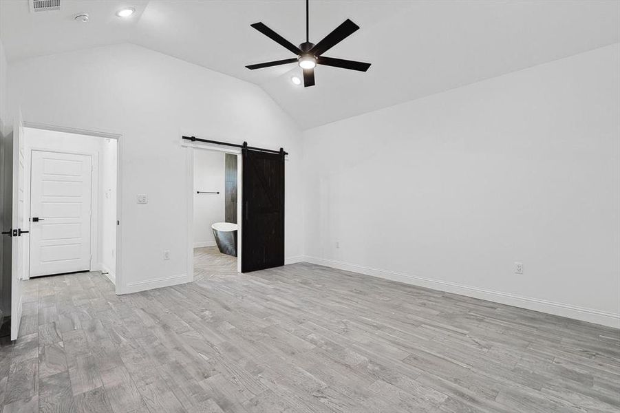 Unfurnished bedroom featuring high vaulted ceiling, ceiling fan, ensuite bath, a barn door, and light hardwood / wood-style flooring