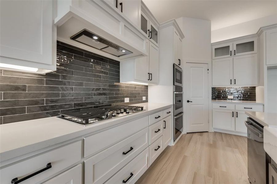 Kitchen with white cabinetry, light hardwood / wood-style flooring, appliances with stainless steel finishes, premium range hood, and tasteful backsplash