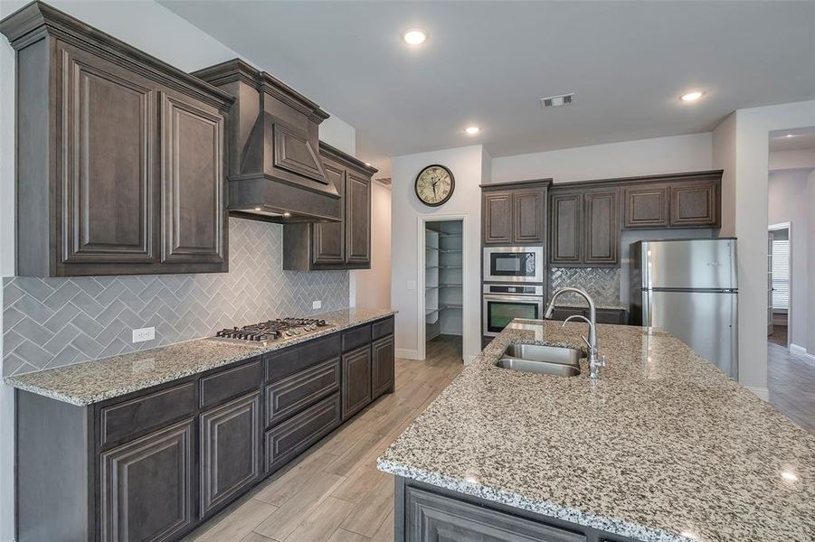 Kitchen with stainless steel appliances, sink, dark brown cabinetry, light wood-type flooring, and premium range hood