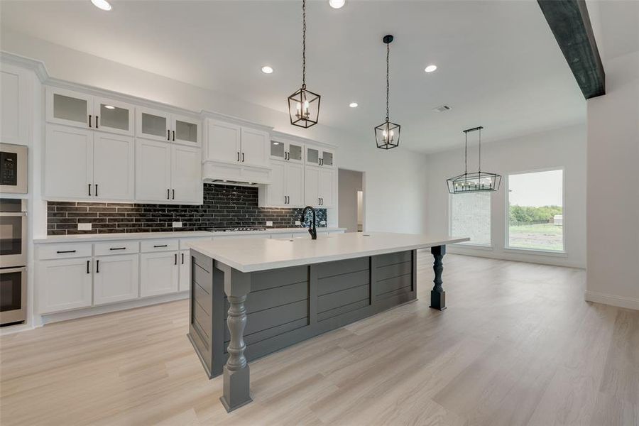 Kitchen with white cabinets, a center island with sink, hanging light fixtures, backsplash, and light hardwood / wood-style floors