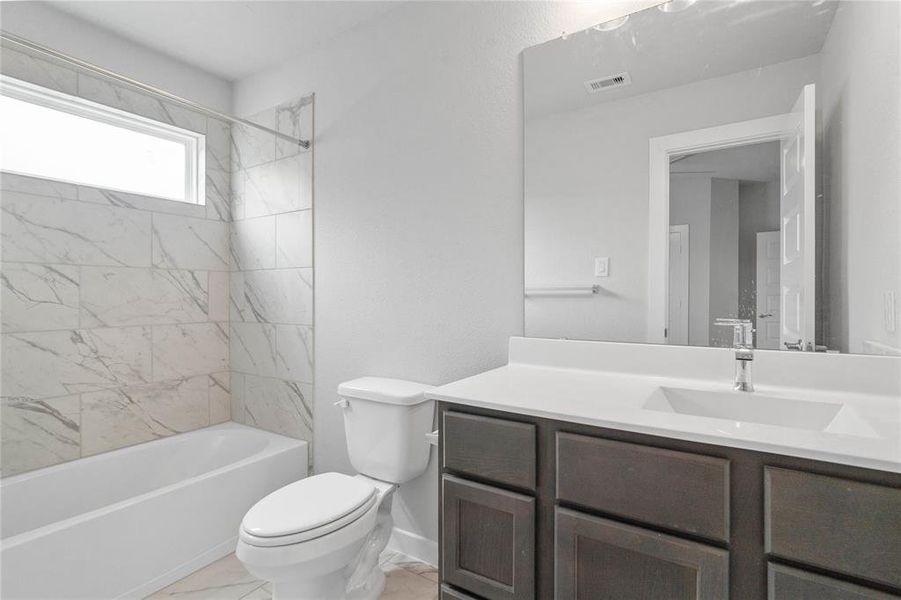 Secondary bath features tile flooring, bath/shower combo with tile surround, wood stained cabinets, beautiful light countertops, mirror, dark, sleek fixtures and modern finishes!