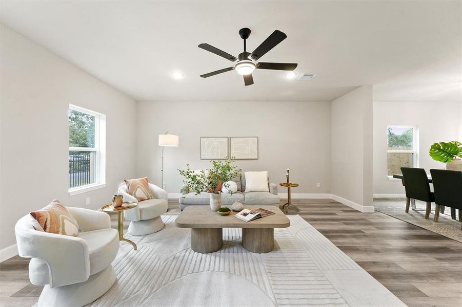 This is a bright, contemporary living space featuring neutral colors, hardwood floors, and modern fixtures. The open floor plan includes a cozy seating area and a dining space with a view of the outdoors through large windows. A ceiling fan adds comfort, and the room's design offers both elegance and functionality.