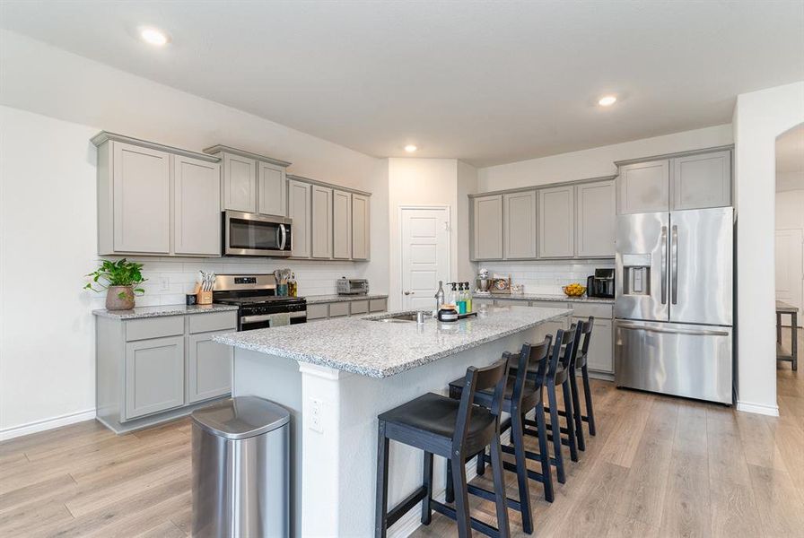 Kitchen featuring decorative backsplash, light hardwood / wood-style flooring, stainless steel appliances, a center island with sink, and light stone countertops