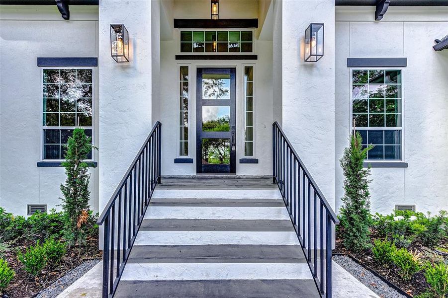 Stairs flanked by wrought iron railing lead to a welcoming front porch. The door features glass inserts and surrounding transom windows allowing in plenty of natural light.