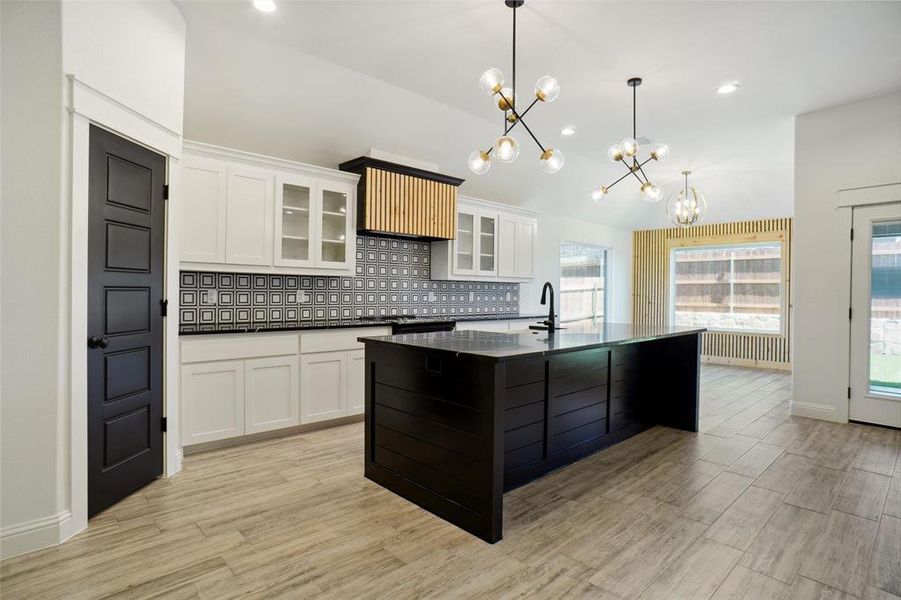 Kitchen with tasteful backsplash, light wood-type flooring, white cabinets, an island with sink, and decorative light fixtures