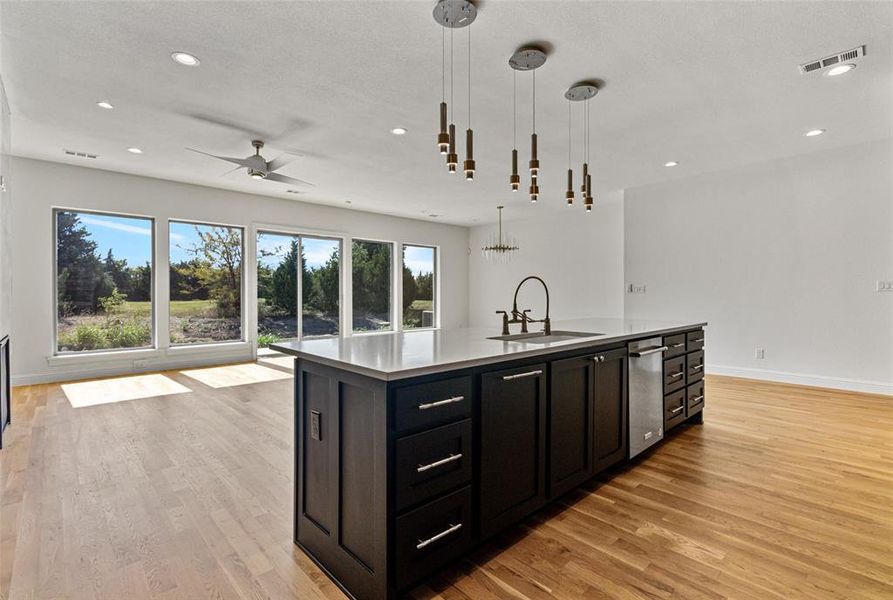 Kitchen featuring sink, light wood-type flooring, an island with sink, ceiling fan with notable chandelier, and hanging light fixtures