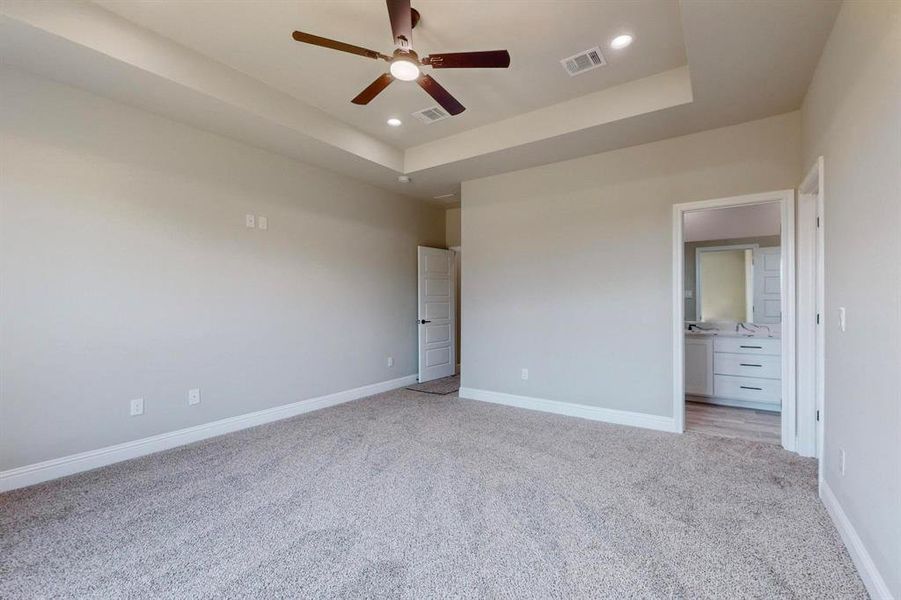 Unfurnished bedroom featuring ceiling fan, light colored carpet, a tray ceiling, and ensuite bath