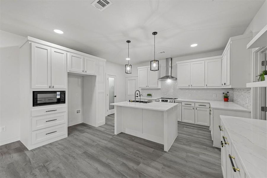 Kitchen featuring wall chimney range hood, light hardwood / wood-style floors, a kitchen island with sink, pendant lighting, and white cabinetry