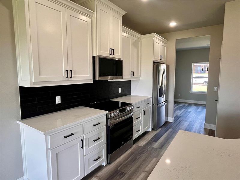 Kitchen with dark wood-type flooring, stainless steel appliances, light stone countertops, backsplash, and white cabinetry