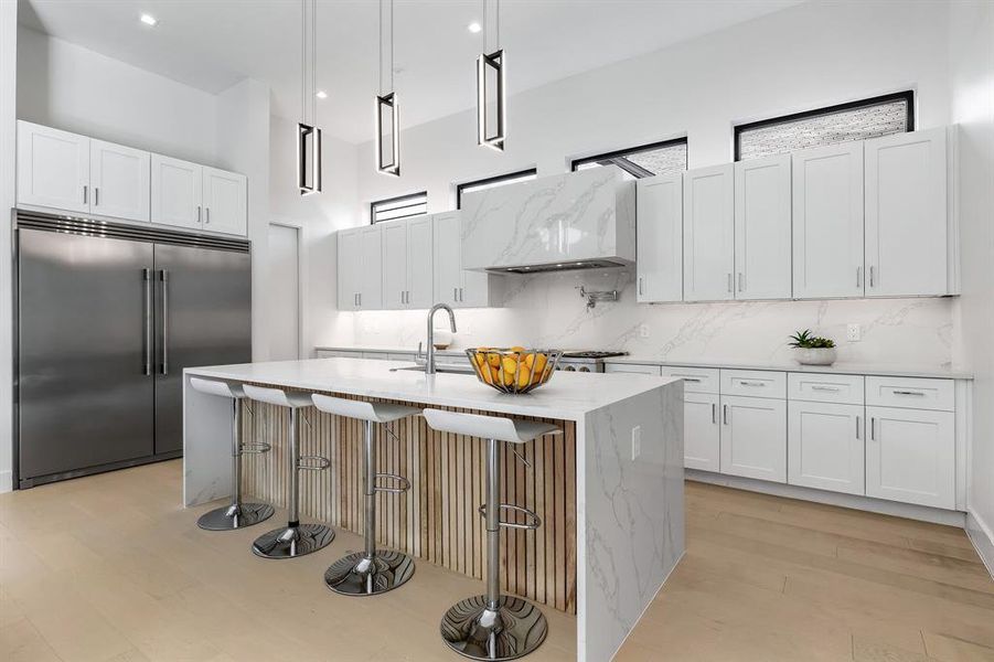 Kitchen with white cabinets, light wood-type flooring, light stone counters, built in fridge, and a kitchen island with sink