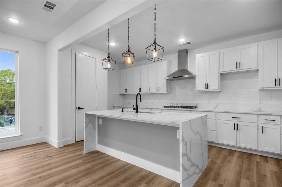 Kitchen with wall chimney range hood, sink, an island with sink, decorative light fixtures, and white cabinets