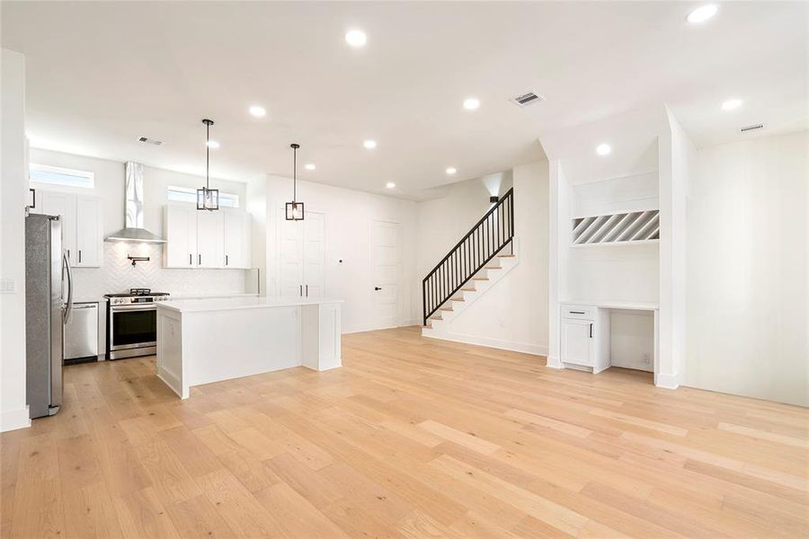 This is a bright, modern open-concept space featuring a kitchen with stainless steel appliances and white cabinetry, a spacious living area with recessed lighting, and hardwood floors throughout. A staircase with a sleek railing leads to the upper level, and there's a built-in desk/ or use as a dry bar under the stairs for convenience.