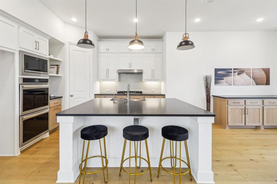 Kitchen featuring light hardwood / wood-style flooring, stainless steel appliances, a center island with sink, backsplash, and hanging light fixtures