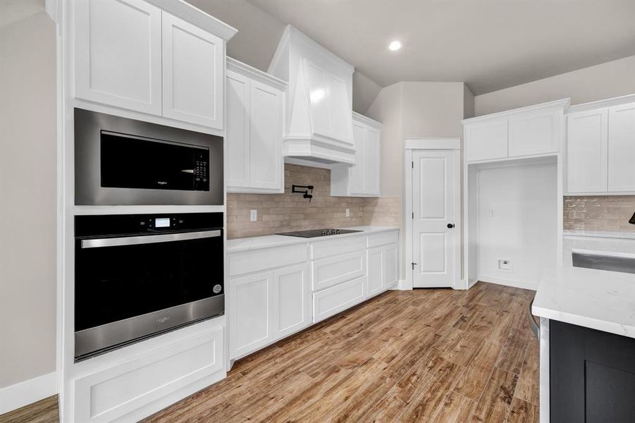 Kitchen featuring backsplash, custom exhaust hood, black appliances, white cabinetry, and light wood-type flooring
