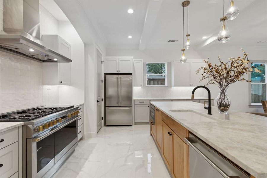 Kitchen featuring premium appliances, decorative light fixtures, wall chimney range hood, white cabinetry, and light stone countertops