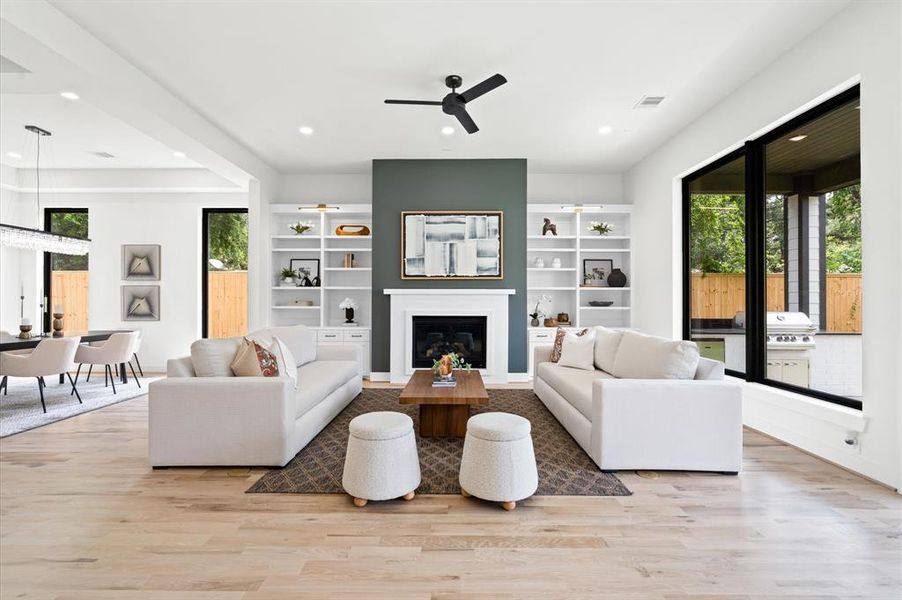 Spacious living room with white built-ins, fireplace, stone mantle; opens to a dining area with large windows, modern decor, and neutral tones.