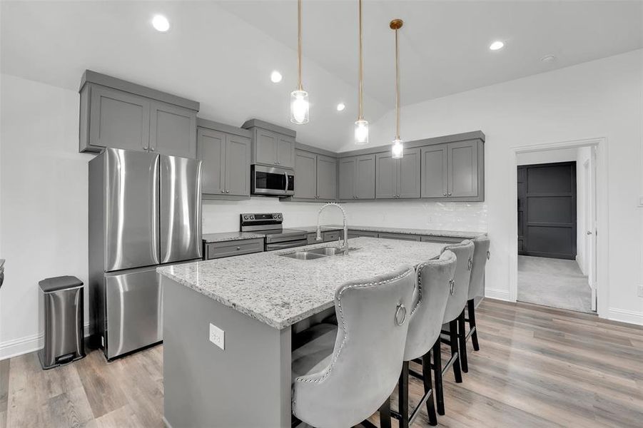 Kitchen with light stone counters, vaulted ceiling, sink, an island with sink, and appliances with stainless steel finishes