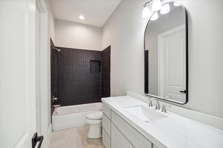 This is a modern bathroom featuring a dark-tiled bathtub with shower, a white toilet, and a vanity with a sink and large mirror. The space has neutral colors and is well-lit.