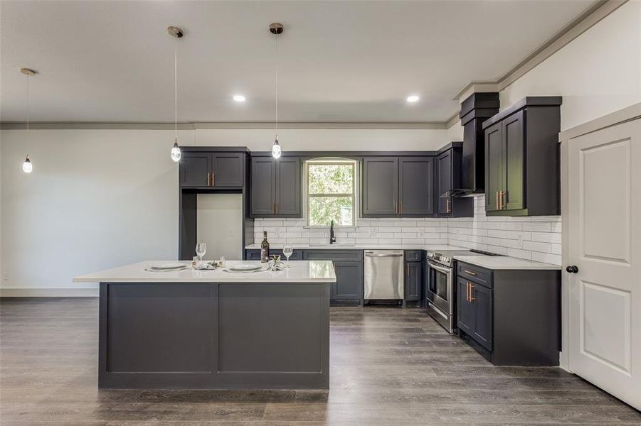 Kitchen featuring stainless steel appliances, a center island with sink, decorative light fixtures, wood-type flooring, and wall chimney exhaust hood