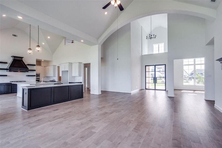 Kitchen featuring light hardwood / wood-style floors, an island with sink, premium range hood, decorative light fixtures, and high vaulted ceiling