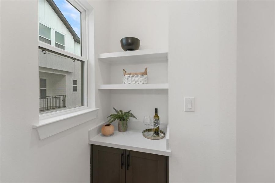 Your modern dry bar area with built-in shelving and a quartz countertop. There's a large window providing natural light, and the space features clean lines and a simple, elegant design.