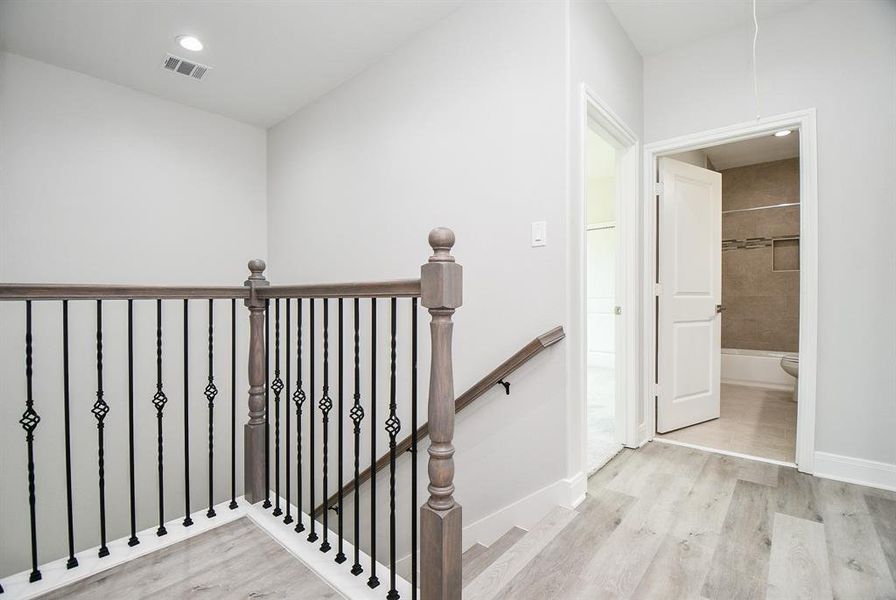 The photo shows a modern, well-lit hallway with elegant wood and wrought iron railings. Neutral colors and contemporary fixtures complement the space, with a glimpse into a bathroom featuring tiled walls.