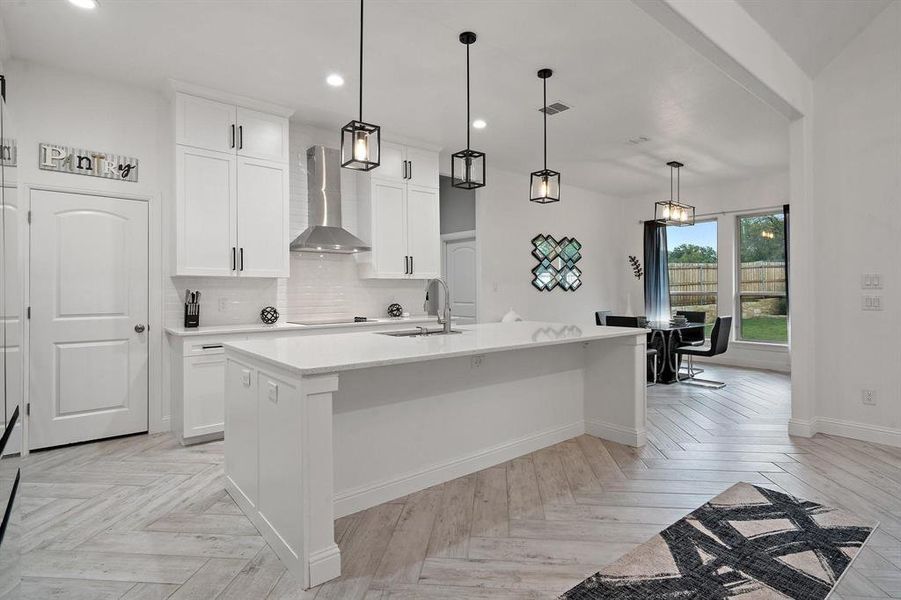 Kitchen featuring tasteful backsplash, white cabinets, hanging light fixtures, wall chimney range hood, and a center island with sink