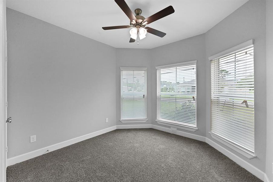 This is a bright, empty room with neutral gray walls, carpeted flooring, three large windows with blinds, and a ceiling fan with lighting. It appears clean and ready for move-in.