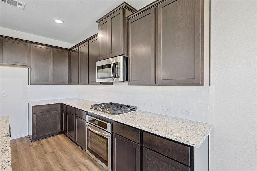 Kitchen with dark brown cabinets, tasteful backsplash, light wood-type flooring, light stone counters, and appliances with stainless steel finishes