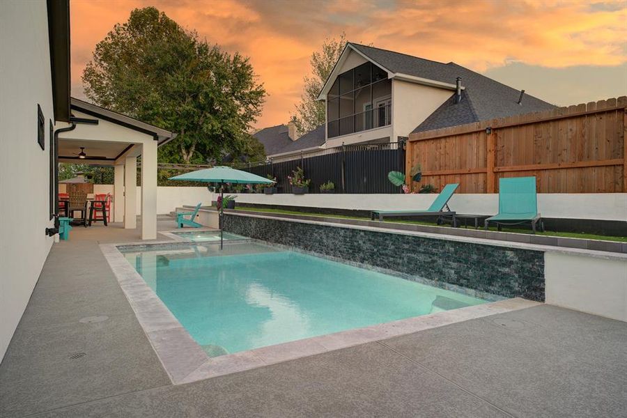 An inviting covered patio area with seating, and a neatly fenced perimeter for privacy. The setting is complemented by a picturesque sunset sky.