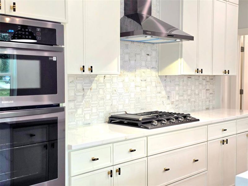 Kitchen featuring white cabinetry, gas stovetop, backsplash, double oven, and wall chimney exhaust hood