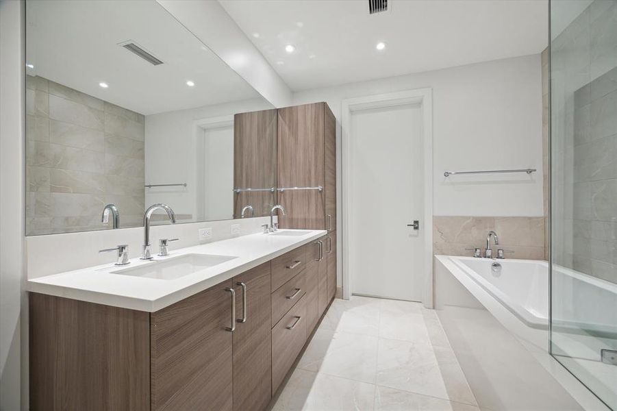 Another view of the primary bathroom featuring a double vanity with a sleek wooden finish cabinetry, a large mirror, and a full-size soaking bathtub. Room has neutral-toned tiles and bright lighting, creating a clean and spacious feel.