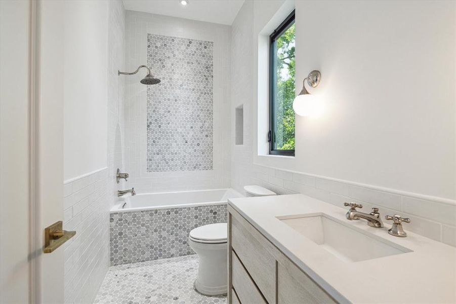 This stylish ensuite bathroom features a tub/shower combo framed by marble penny round floor and wall tile, adding a touch of luxury. Quartz countertops, designer polished nickel sconces, and a stunning bleached oak vanity complete the sophisticated design.