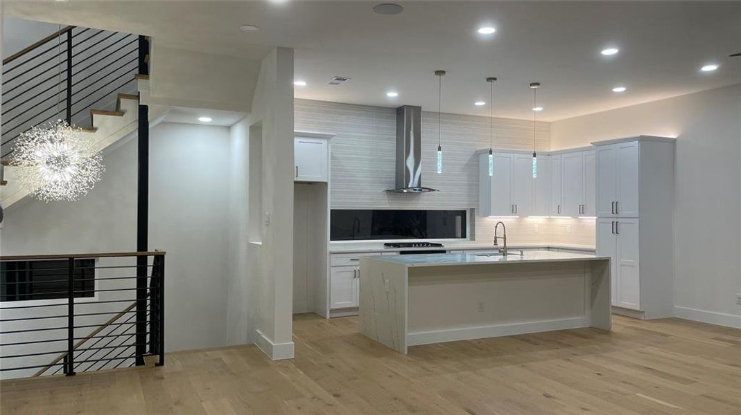 Kitchen with light hardwood / wood-style floors, an island with sink, white cabinets, hanging light fixtures, and wall chimney range hood