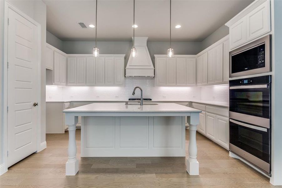 Kitchen featuring custom range hood, stainless steel appliances, backsplash, light wood-type flooring, and white cabinets