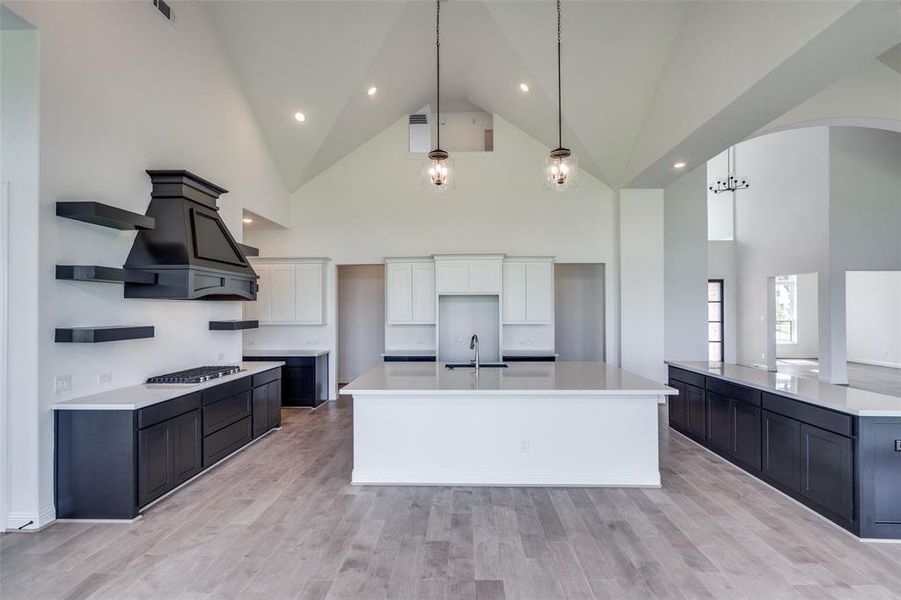 Kitchen featuring an island with sink, decorative light fixtures, light wood-type flooring, high vaulted ceiling, and white cabinets