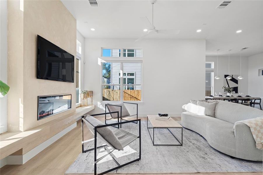 This light-filled living space boasts soaring ceilings and oversized windows that flood the room with natural light. The sleek fireplace, framed by a Roman Clay accent wall, becomes an instant focal point, while the seamless flow into the dining area creates the perfect environment for both relaxation and entertaining. Clean lines, contemporary finishes, and an open layout define this inviting and versatile space.