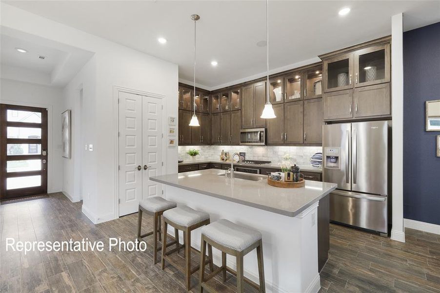 Kitchen featuring backsplash, stainless steel appliances, an island with sink, dark brown cabinetry, and pendant lighting