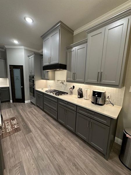 Kitchen with backsplash, gray cabinetry, double wall oven, crown molding, and wood-type flooring