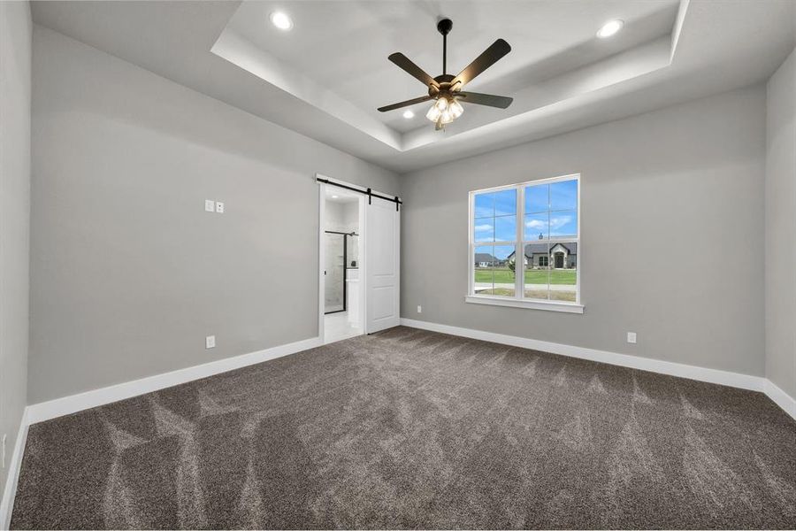 Primary bedroom with a barn door to restroom, a tray ceiling, carpet flooring,