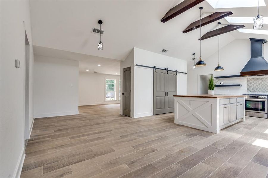 Kitchen with custom exhaust hood, butcher block counters covering a massive island, beamed ceiling, a barn door, and high end flooring facing the large dining area.