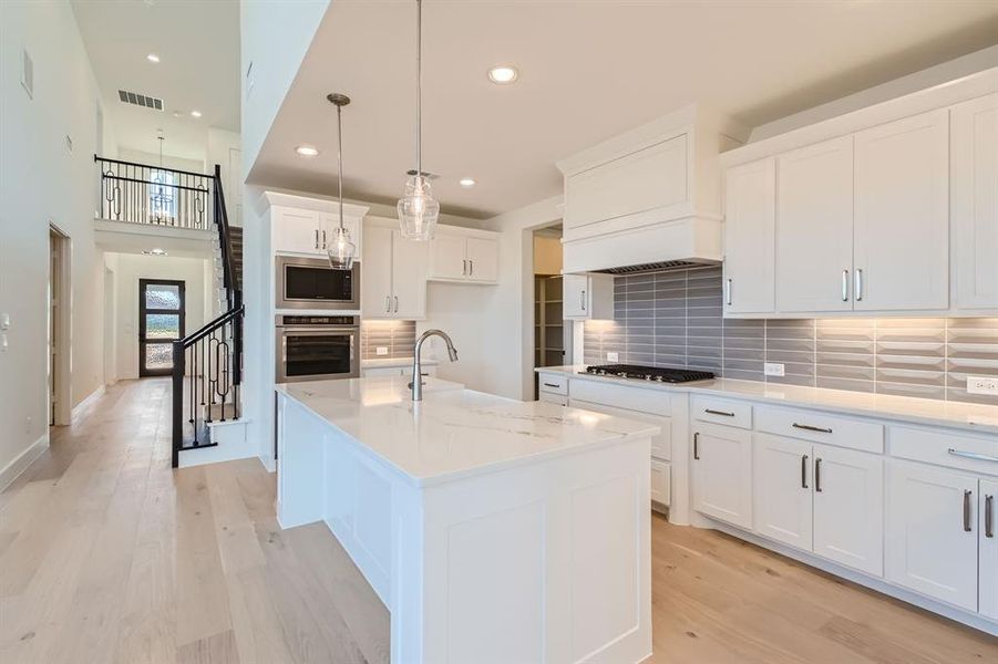 Kitchen with an island with sink, stainless steel appliances, decorative light fixtures, white cabinets, and light hardwood / wood-style floors